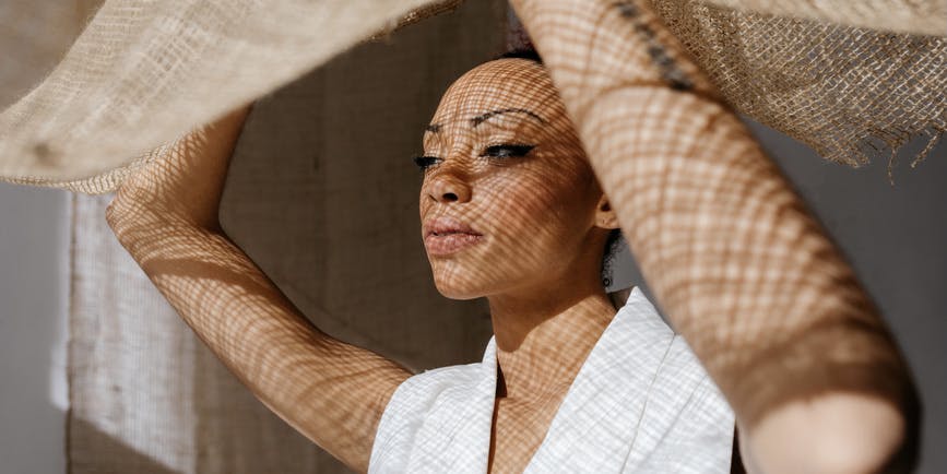 Pensive black lady with makeup and bare shoulders looking away while raising brown cloth in daylight and having light spots on body.